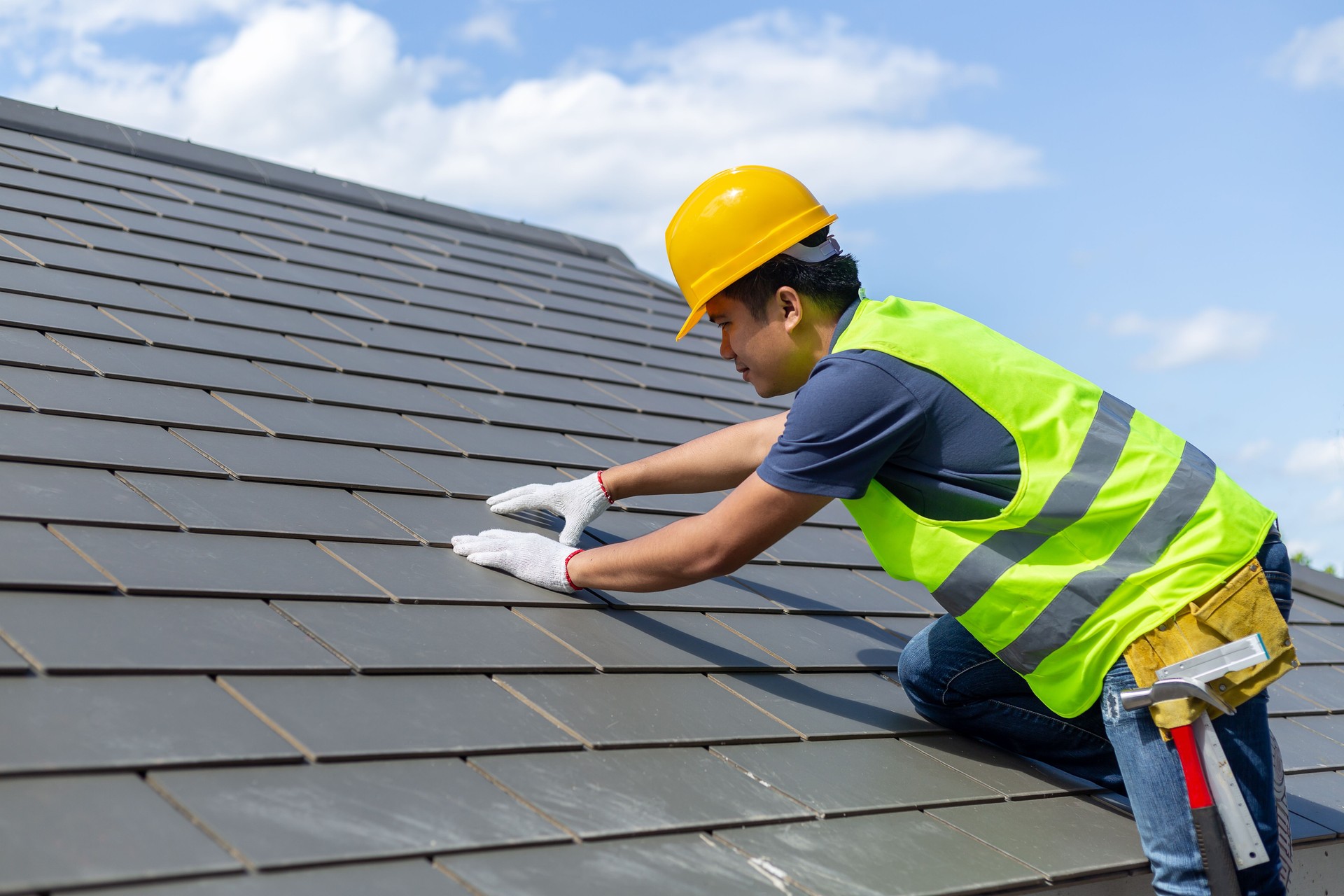 Roof repair, worker with white gloves replacing gray tiles or shingles on house with blue sky as background and copy space, Roofing - construction worker standing on a roof covering it with tiles.